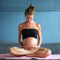 a pregnant woman sitting on a yoga mat in front of a blue wall and holding her belly