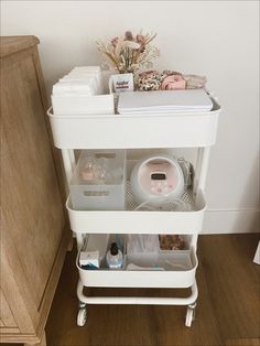 a white cart with many items in it on top of a wooden floor next to a dresser