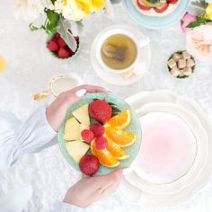 a person holding a bowl of fruit on top of a table with plates and cups