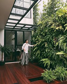 a person standing on a deck in front of a green wall and some plants with their arms out