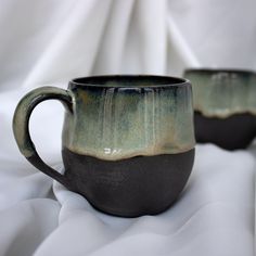 two coffee mugs sitting on top of a white cloth covered tablecloth, one is green and the other is black