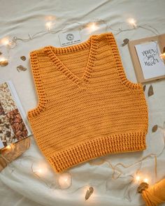 an orange knitted sweater sits on a bed next to a book and some string lights