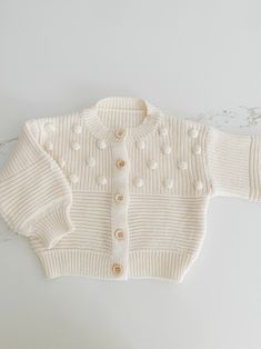 Soft and cozy, this stunning knit cardigan is the perfect addition to your little ones closet. Tiny Pom Pom appliqués all over. Textured pattern with front buttons. Playful Knit Cardigan For Winter, Playful Knit Cardigan For Fall, Playful Knit Long Sleeve Outerwear, Playful Cotton Knitted Cardigan, Playful Knitted Cotton Cardigan, Cute Cream Cotton Cardigan, Playful Long Sleeve Knit Cardigan, Cute Long Sleeve Knit Cardigan, Cute Knitted Cotton Cardigan