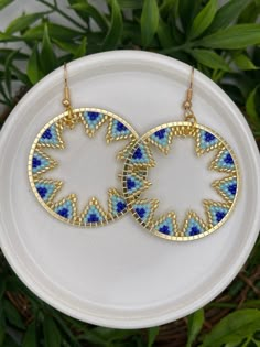 two gold and blue earrings sitting on top of a white plate next to green leaves