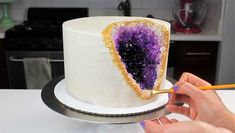 a woman is decorating a white cake with purple and gold decorations on it while holding a pencil in her hand