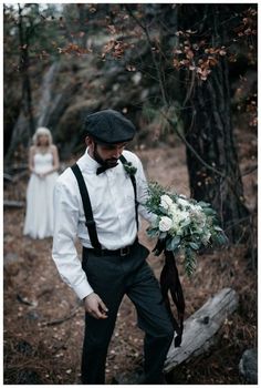 a man wearing a hat and suspenders holding a bridal bouquet in the woods