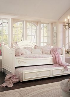 a white daybed with pink pillows and blankets on it in front of two windows