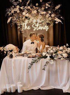 a bride and groom kissing in front of a table with white flowers, greenery and hanging lights