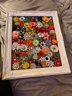 a white frame with many colorful flowers in it on a gray bed sheet and someone's hand holding the frame