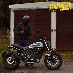 a man standing next to a motorcycle in front of a garage door with the word scramble written on it