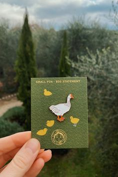 a hand holding up a small card with a duck on it's back and some trees in the background