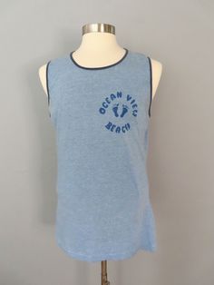 Vintage sleeveless t-shirt. Blue heather with dark blue trim around the neck and sleeves. Graphic on right breast Ocean View Beach with foot prints.  MEASUREMENTS--Taken with garment laying flat, in inches, doubled: Shoulder: 15.5" Chest: 44" Waist: 38" Overall length from shoulder to hem: 29.5" Label/ Era: Hanes | 1980 Fabric: Cotton, polyester Condition: Great condition. Cleaned and ready to wear.  ★ Shop entire shop here: https://www.etsy.com/shop/retrosuzysvintage Instagram | retrosuzysvintage Facebook | retrosuzysvintage Vintage Beach Shirt With Vintage Print, Vintage Summer Beach T-shirt, Blue Vintage Beach Shirt, Vintage Graphic Print Beach T-shirt, 90s Cotton T-shirt For Beach, Ocean Views Beach, Sleeveless Tshirt, Ocean View, Overalls