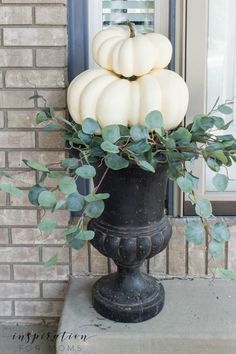 white pumpkins are stacked on top of a black urn with greenery in front of a brick building