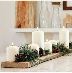 some white candles are sitting on a wooden board with pine cones and greenery around them