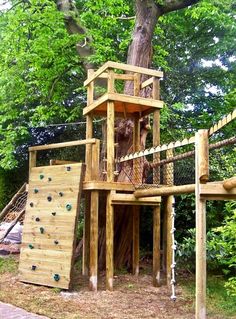 a wooden play structure in the middle of a park with climbing ropes and a tree