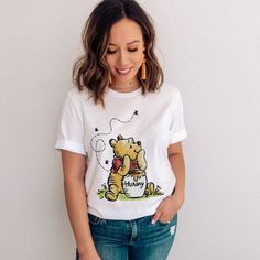 Step back into the whimsical world of Winnie the Pooh with our "Winnie the Pooh with the Honey Pot" T-Shirt. This tee features a classic retro design with a large print of Pooh bear and his iconic honey pot, complete with the amusing detail of three flies buzzing around. A perfect homage to A.A. Milne's beloved character, this tee is an adorable and cute gift for fans of all ages. Celebrate the nostalgia of childhood and share the joy of Pooh's sweet adventures with this delightful tee. Bella Ca Cute White T-shirt With Bear Design, Cute Cartoon Print T-shirt For Gift, Cute Cotton T-shirt With Bear Design, Cute Bear Print Crew Neck T-shirt, Cute Cartoon Print T-shirt As Gift, Cute Short Sleeve T-shirt With Bear Print, The Honey Pot, Pooh Bear, Honey Pot