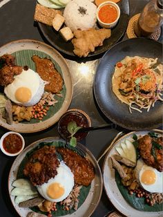 four plates filled with food on top of a black countertop next to cups and sauces