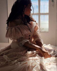 a woman sitting on top of a bed next to a window in a white dress