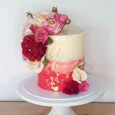 a white cake with pink and red flowers on top, sitting on a stand in front of a wall