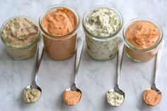 four jars filled with different types of dips and spoons on a marble surface