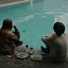 two people sitting on the edge of a swimming pool with plates and wine glasses in front of them