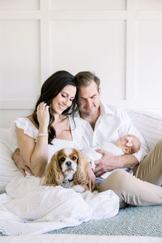 a man and woman sitting on a bed with a dog in their lap while holding a baby