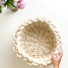 a hand holding a woven basket next to pink flowers and a potted plant on the table