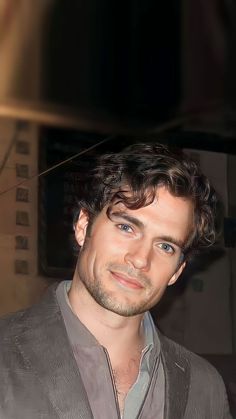 a man with curly hair and blue eyes smiles at the camera while wearing a gray jacket