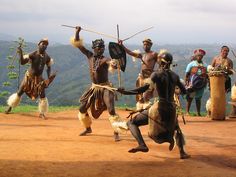 some people are dancing on a dirt road with mountains in the background and one person is holding two sticks