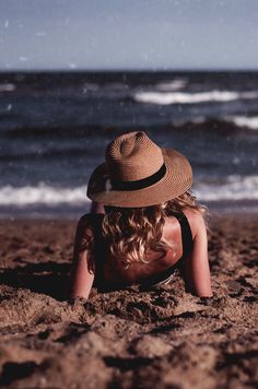 a woman laying on top of a sandy beach next to the ocean in a hat