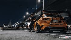 a man standing next to an orange car on the side of a road at night