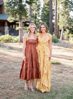 two women standing next to each other in front of some trees and grass with one wearing a brown dress