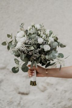 a person holding a bouquet of flowers and greenery in their hand on the beach