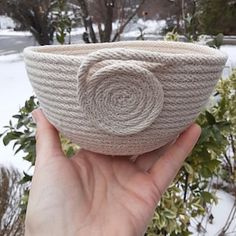 a hand holding up a knitted bowl with a flower on the side in front of snow covered trees