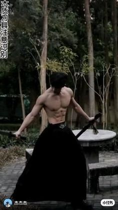 a shirtless man holding two swords in front of his face and back with trees behind him