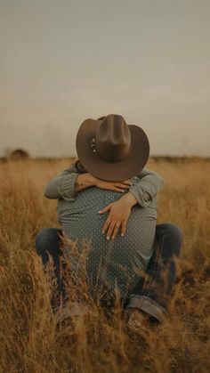 a woman sitting in the middle of a field wearing a cowboy hat and hugging her stomach