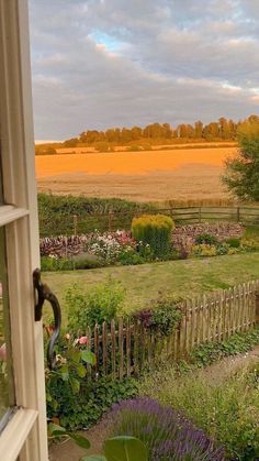 an open window looking out onto a garden and field in the distance with trees, shrubs, and flowers