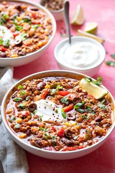 two bowls filled with chili, beans and sour cream on top of a pink table