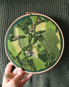 someone is holding up a cross stitch pattern in front of a green background with trees and houses