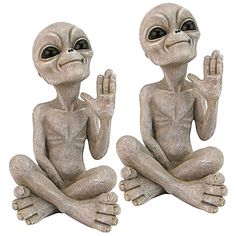 two alien statues sitting next to each other on a white background, one holding his hands in the air