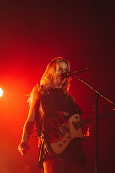 a woman in black dress holding a guitar on stage with red light coming from behind her