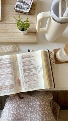 an open book sitting on top of a wooden table next to a cup and plant