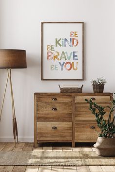 a room with a dresser, lamp and painting on the wall above it that says be kind of brave be you