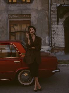 a woman standing next to a red car talking on her cell phone while wearing a suit