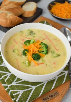 a bowl of broccoli and cheese soup on a cutting board with bread in the background