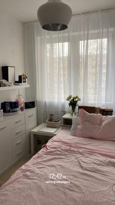 a bed with pink sheets and pillows in front of a window next to a white dresser