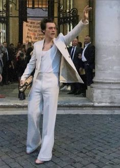 a man in white suit and tie standing on the street with his hand raised up