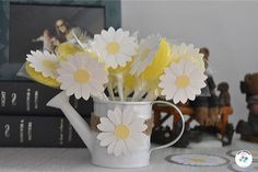 a white watering can filled with daisies on top of a table next to books