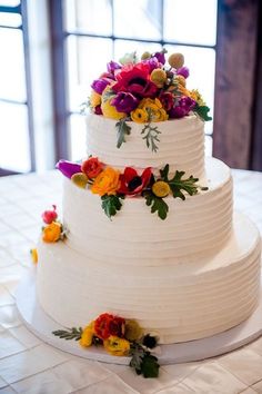 a three tiered white cake with colorful flowers on the top and bottom, sitting on a table