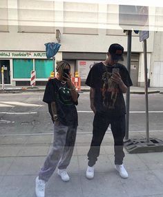 two young men standing next to each other on the sidewalk while looking at their cell phones